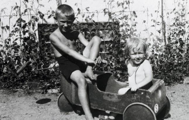 A black-and-white archive photograph of two young children in a garden, one sitting on the edge of a wooden toy car and the other sitting inside it.