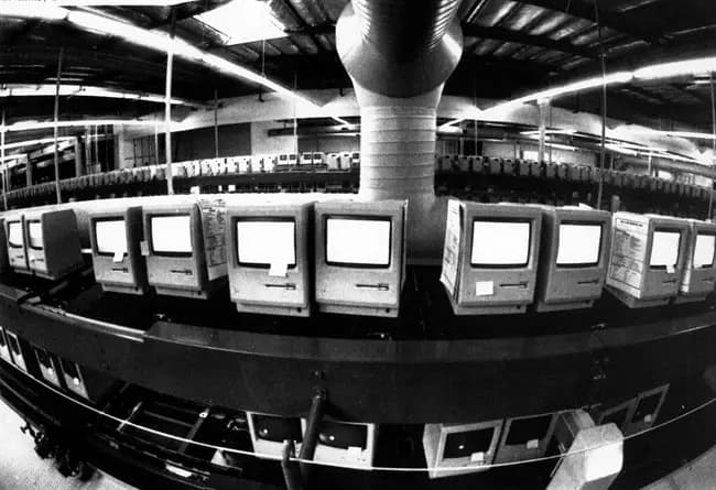 Rows of early Macintosh computers moving along a factory production line under long fluorescent lights.